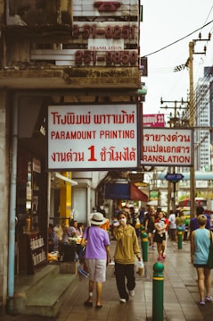 A busy urban street scene featuring people walking along a sidewalk lined with shops and signs in both English and Thai languages. The atmosphere appears lively with a mix of individuals casually strolling or engaged in conversation. Visible signs include those for a printing shop and translation services. Various utility poles and a cityscape can be seen in the background.