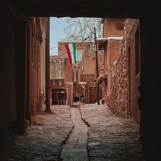 A narrow cobblestone alleyway leads to a small courtyard with traditional brown buildings. An Iranian flag is prominently displayed, hanging from a pole. The architecture is rustic and historic, with rough stonework and wooden elements.