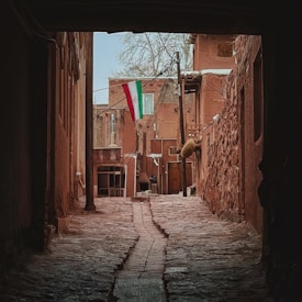A narrow cobblestone alleyway leads to a small courtyard with traditional brown buildings. An Iranian flag is prominently displayed, hanging from a pole. The architecture is rustic and historic, with rough stonework and wooden elements.