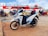 A white and blue Honda scooter is parked on a concrete surface near a dock. Several other motorcycles and riders wearing helmets are visible in the background, along with a large passenger ferry featuring red and white colors docked next to them. The sky is partly cloudy.
