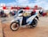 A white and blue Honda scooter is parked on a concrete surface near a dock. Several other motorcycles and riders wearing helmets are visible in the background, along with a large passenger ferry featuring red and white colors docked next to them. The sky is partly cloudy.