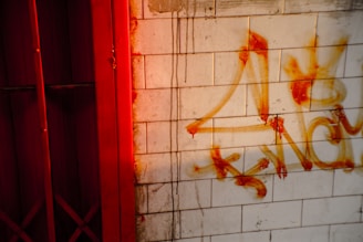 Graffiti sprayed in orange paint on a tiled wall, with streaks and drips. A red metal gate or door is partially visible to the left, with shadows and light giving a moody contrast.