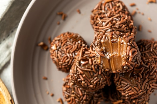 A close-up view of chocolate-covered candies arranged in a pile on a plate. The candies are coated with chocolate sprinkles, and some pieces reveal the smooth, creamy interior filling.
