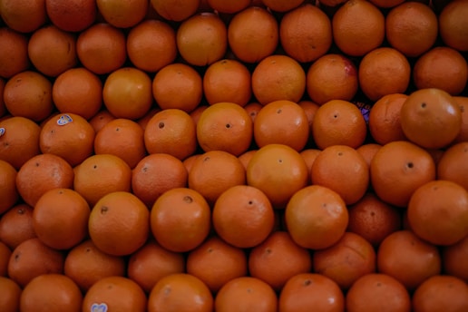 A large pile of fresh oranges is closely packed together, showcasing their vibrant orange color and smooth texture. Some oranges have small stickers on them, indicating they may be for sale at a market or grocery store.