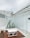 A minimalist outdoor seating area with white metal-framed glass panels and a wooden floor. The space includes white geometric tables and a row of tall, potted cacti. The overall design is modern and sleek, with a focus on angular lines.