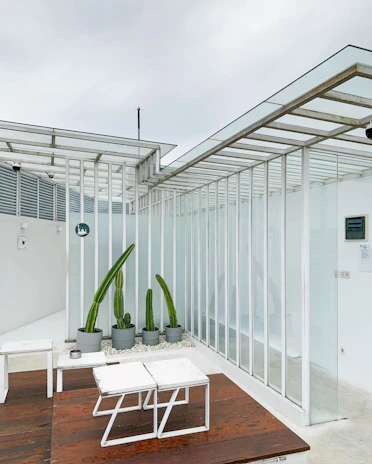 A daytime view of a minimalist outdoor lounge area framed by a sleek metal pergola and lush greenery.