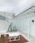 A minimalist outdoor seating area with white metal-framed glass panels and a wooden floor. The space includes white geometric tables and a row of tall, potted cacti. The overall design is modern and sleek, with a focus on angular lines.