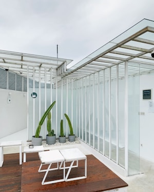 A minimalist outdoor seating area with white metal-framed glass panels and a wooden floor. The space includes white geometric tables and a row of tall, potted cacti. The overall design is modern and sleek, with a focus on angular lines.