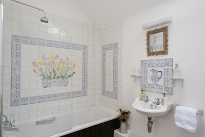 A bathroom with white tiled walls, featuring a floral design with tulips and daffodils above a bathtub. A vintage-style mirror with a decorative frame hangs above a small white sink that has blue patterned tile design. A towel hangs nearby, and various toiletries are neatly arranged.