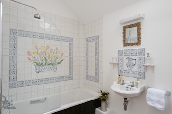A bathroom with white tiled walls, featuring a floral design with tulips and daffodils above a bathtub. A vintage-style mirror with a decorative frame hangs above a small white sink that has blue patterned tile design. A towel hangs nearby, and various toiletries are neatly arranged.