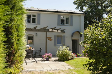 A charming two-story house with light gray shutters and a wooden pergola at the entrance, surrounded by lush greenery. A small patio is visible with a table and chairs, adorned with potted flowers and plants. The scene is sunlit, indicating a clear, pleasant day.