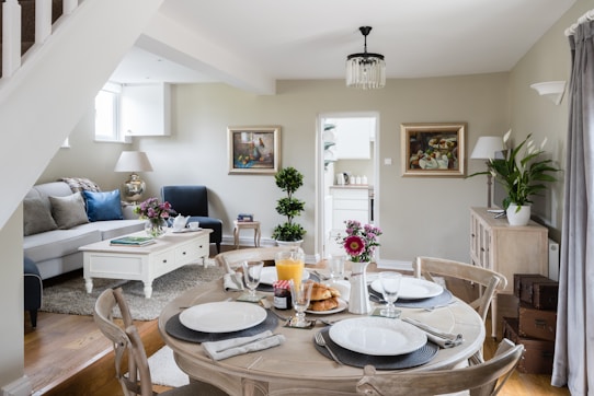 A cozy and well-decorated living and dining area with a round wooden dining table set for a meal. There are four place settings with plates, glasses, napkins, and cutlery. In the center of the table, there is orange juice, croissants, and a vase with flowers. The background features a living room with a white coffee table, blue and white sofa, armchair, and two framed paintings on the wall. A staircase and indoor plants are also visible, along with a glimpse into an adjoining kitchen.