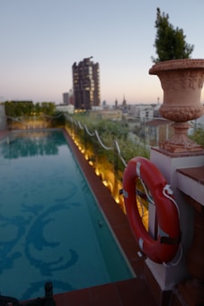 A rooftop swimming pool surrounded by glass barriers and decorated with small shrubs and yellow accent lighting. In the foreground, there's a red lifebuoy attached to a white column with a decorative flower pot. The background features a cityscape with tall buildings under a clear evening sky.