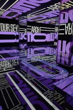 A visually striking room filled with mirrored surfaces reflecting large, bold text in purple and white. The text covers the walls, ceiling, and floor, creating an immersive and repetitive visual effect. The reflections and lighting add a sense of infinity and depth to the space.