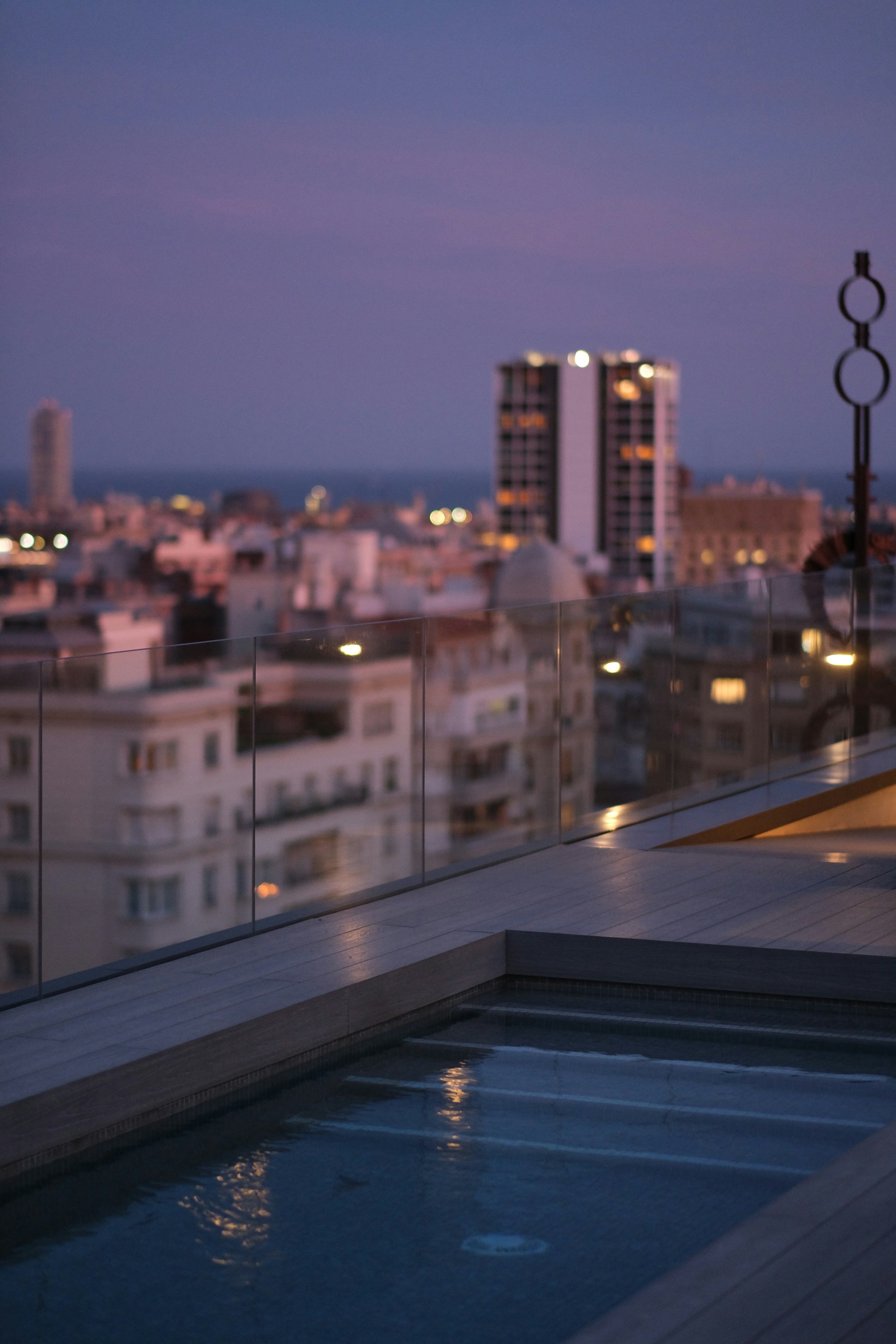 A rooftop pool is surrounded by a clear glass barrier, overlooking a cityscape at dusk. The sky is transitioning from blue to a soft purple, and city lights are beginning to twinkle. A tall building prominently stands in the background, contrasting with shorter surrounding structures.
