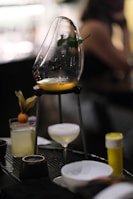 A sophisticated cocktail setup featuring a wooden or metal stand holding a curved glass vessel with a small portion of amber liquid at the base and a sprig of herbs inside. Below, there is a coupe glass with a foamy white drink, and to the side, a highball glass garnished with a small round fruit. The background is blurred, suggesting a bar or restaurant environment.