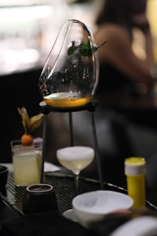 A sophisticated cocktail setup featuring a wooden or metal stand holding a curved glass vessel with a small portion of amber liquid at the base and a sprig of herbs inside. Below, there is a coupe glass with a foamy white drink, and to the side, a highball glass garnished with a small round fruit. The background is blurred, suggesting a bar or restaurant environment.