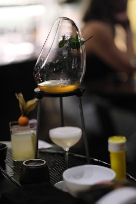 A sophisticated cocktail setup featuring a wooden or metal stand holding a curved glass vessel with a small portion of amber liquid at the base and a sprig of herbs inside. Below, there is a coupe glass with a foamy white drink, and to the side, a highball glass garnished with a small round fruit. The background is blurred, suggesting a bar or restaurant environment.
