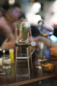 A close-up shot of a glass container with herbs inside, placed on a metal stand on a table. The background shows blurred figures in casual attire, possibly engaged in conversation. Various glasses and a bowl of snacks are also present on the table, creating an ambiance of social interaction.