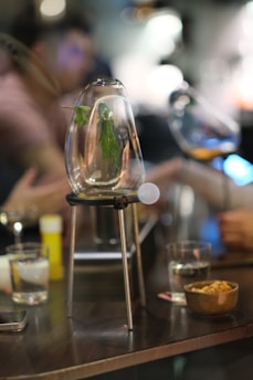 A close-up shot of a glass container with herbs inside, placed on a metal stand on a table. The background shows blurred figures in casual attire, possibly engaged in conversation. Various glasses and a bowl of snacks are also present on the table, creating an ambiance of social interaction.