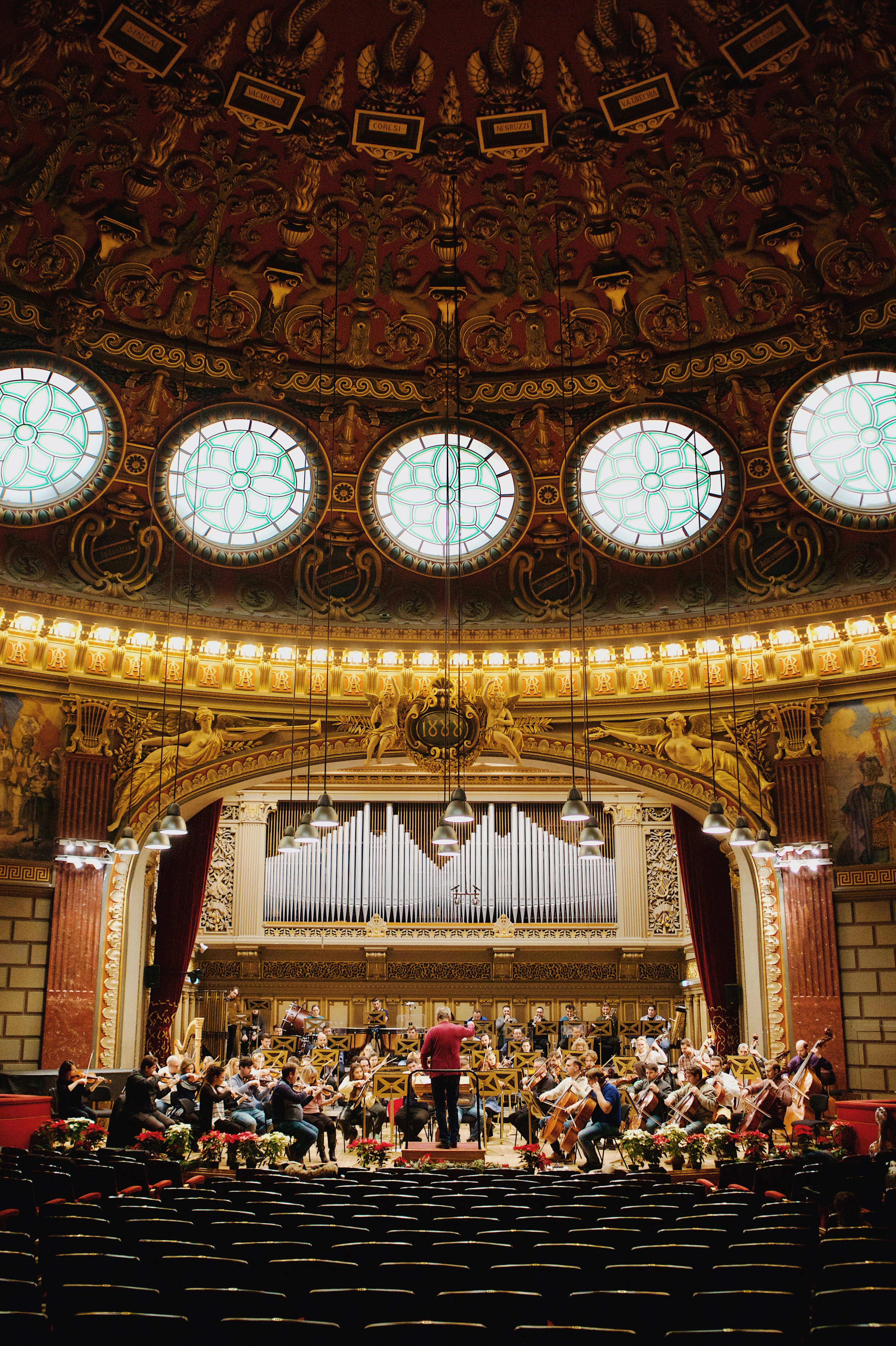 The Grand Hall of the Romanian Athenaeum in Bucharest seen from the empty stalls, an orchestra rehearsing beneath the organ pipes while the gilded dome and its ring of circular stained-glass windows glow overhead.