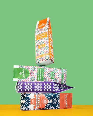 Four colorful coffee bags are stacked in a balanced manner against a green background. The top bag is orange with a vibrant geometric pattern. Below it, a green bag, a purple bag, and a blue bag with intricate designs and branding details are visible.