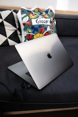 A sleek laptop is placed on a dark couch with geometric-patterned pillows and a colorful cushion featuring floral designs and the word 'Glocal' on it.
