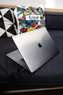 A sleek laptop is placed on a dark couch with geometric-patterned pillows and a colorful cushion featuring floral designs and the word 'Glocal' on it.