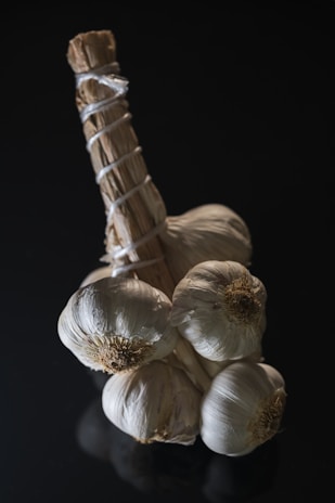 A clustered bunch of garlic bulbs bound together with string against a dark, minimalist background. The garlic has a natural, rustic appearance with visible papery skins and subtle shadows accentuating its texture.