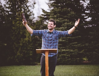 A man wearing a plaid shirt stands outdoors with his arms raised, holding a book in one hand. He is smiling and appears to be speaking or delivering a message. In front of him is a wooden lectern, and behind him are evergreen trees and grass.