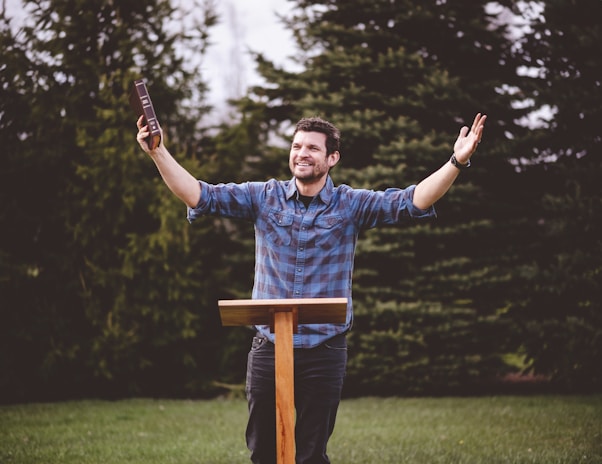 A man wearing a plaid shirt stands outdoors with his arms raised, holding a book in one hand. He is smiling and appears to be speaking or delivering a message. In front of him is a wooden lectern, and behind him are evergreen trees and grass.