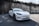 A sleek white electric car is parked on a paved surface with a high rustic wall in the background, partially covered by overgrown greenery and casting shadows on the ground.