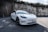 A sleek white electric car is parked on a paved surface with a high rustic wall in the background, partially covered by overgrown greenery and casting shadows on the ground.