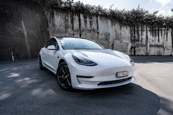 A sleek white electric car is parked on a paved surface with a high rustic wall in the background, partially covered by overgrown greenery and casting shadows on the ground.