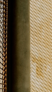 Geometric patterns created by shadows cast through a mesh grid onto a textured surface. The image features a strong play of light and shadow, with a contrast between a dark solid object and a brightly lit area.