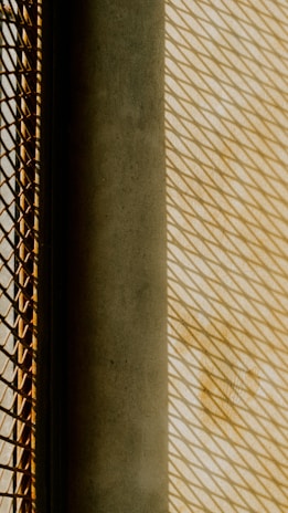 Geometric patterns created by shadows cast through a mesh grid onto a textured surface. The image features a strong play of light and shadow, with a contrast between a dark solid object and a brightly lit area.
