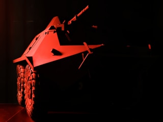 A heavily shadowed image featuring a military armored vehicle with prominent wheels and angular body design. The vehicle is illuminated with a red light, creating a dramatic and intense atmosphere. The details of the environment are obscured by darkness.