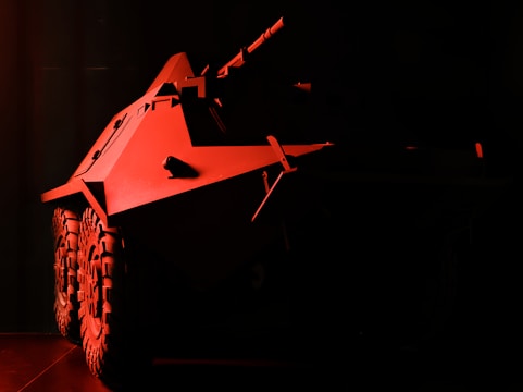 A heavily shadowed image featuring a military armored vehicle with prominent wheels and angular body design. The vehicle is illuminated with a red light, creating a dramatic and intense atmosphere. The details of the environment are obscured by darkness.