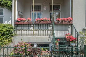 A charming small balcony with a series of vibrant red and pink flowers in wooden planters. The balcony features a decorative metal railing and has a few stairs leading down to a garden area. The facade of the building is painted in light colors, complementing the greenery and flowers.