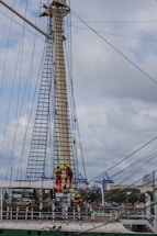 Several workers wearing bright safety gear are climbing and working on a tall ship's mast with rigging lines and ladders. The background features industrial cranes and buildings under a cloudy sky.