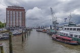 A dock area features several moored boats along a body of water, with a tall red-brick building to the left. A large, old-fashioned crane is situated near the waterway, and cloudy skies loom overhead, creating a slightly overcast atmosphere.