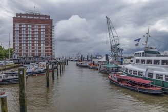 A dock area features several moored boats along a body of water, with a tall red-brick building to the left. A large, old-fashioned crane is situated near the waterway, and cloudy skies loom overhead, creating a slightly overcast atmosphere.