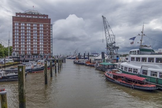 A dock area features several moored boats along a body of water, with a tall red-brick building to the left. A large, old-fashioned crane is situated near the waterway, and cloudy skies loom overhead, creating a slightly overcast atmosphere.