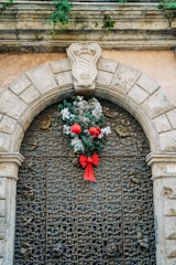 An ornate, wrought-iron gate is framed by a stone archway with intricate detailing. Above the gate, a decorative stone crest adds historical character. A festive Christmas wreath, adorned with red ribbons and ornaments, hangs prominently on the gate. The surrounding stonework is weathered, with patches of greenery suggesting age and natural beauty.