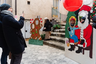 A group of people enjoy a festive scene on a cobblestone street. One person is capturing a photo using a smartphone. Two cardboard cutouts are present: one featuring a gingerbread figure and another depicting Santa Claus with a child. Two people have placed their faces in the cutouts to pose for a picture. Nearby, a large red balloon and a decorated Christmas tree contribute to the holiday theme.