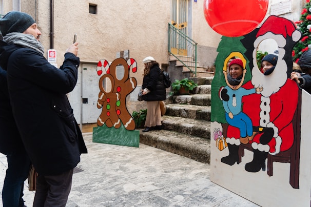 A group of people enjoy a festive scene on a cobblestone street. One person is capturing a photo using a smartphone. Two cardboard cutouts are present: one featuring a gingerbread figure and another depicting Santa Claus with a child. Two people have placed their faces in the cutouts to pose for a picture. Nearby, a large red balloon and a decorated Christmas tree contribute to the holiday theme.