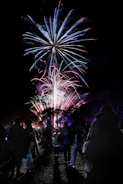A large display of colorful fireworks lights up the night sky. Below, silhouetted figures of people stand together in an open area, watching the spectacle above. The crowd creates a sense of community and celebration.