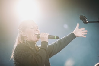A person passionately singing into a microphone with an outstretched arm. The bright light source behind creates a dramatic and ethereal atmosphere.