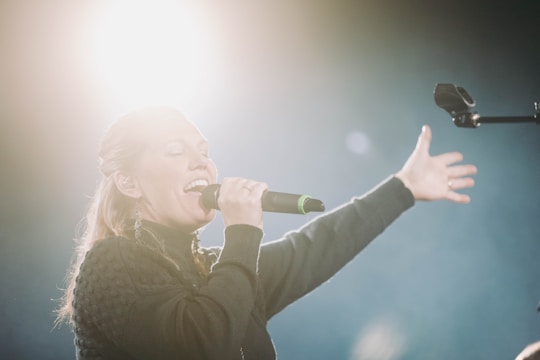A person passionately singing into a microphone with an outstretched arm. The bright light source behind creates a dramatic and ethereal atmosphere.