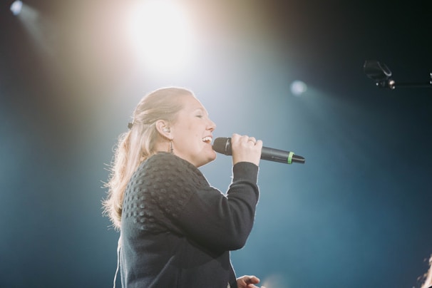 A person is singing into a microphone, with bright stage lights illuminating the background. The individual appears to be performing on a stage, wearing a dark long-sleeve sweater. The focus is on the singer against a backdrop of diffused light, creating a dramatic effect.